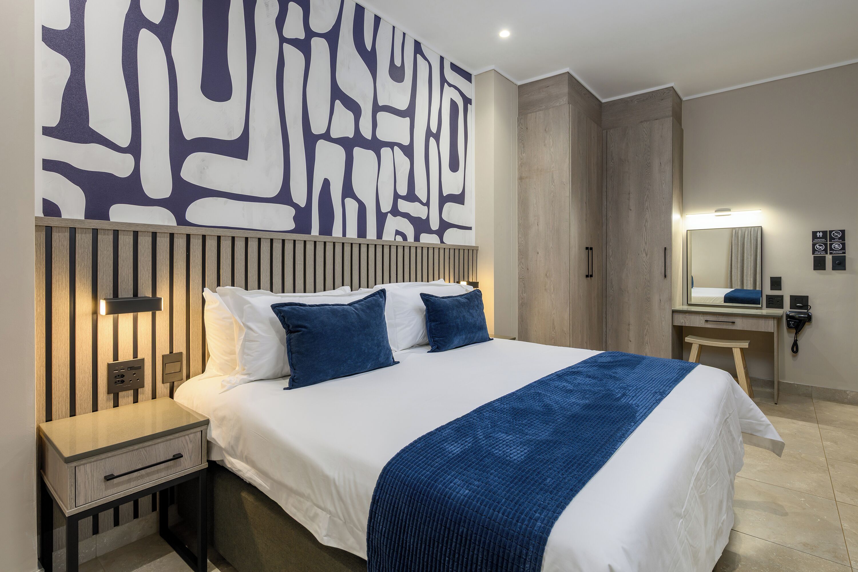 Ein Hotelzimmer mit Bett, Nachttisch, Wandbildern, einem Spiegel und einer Wandverkleidung aus Holz.