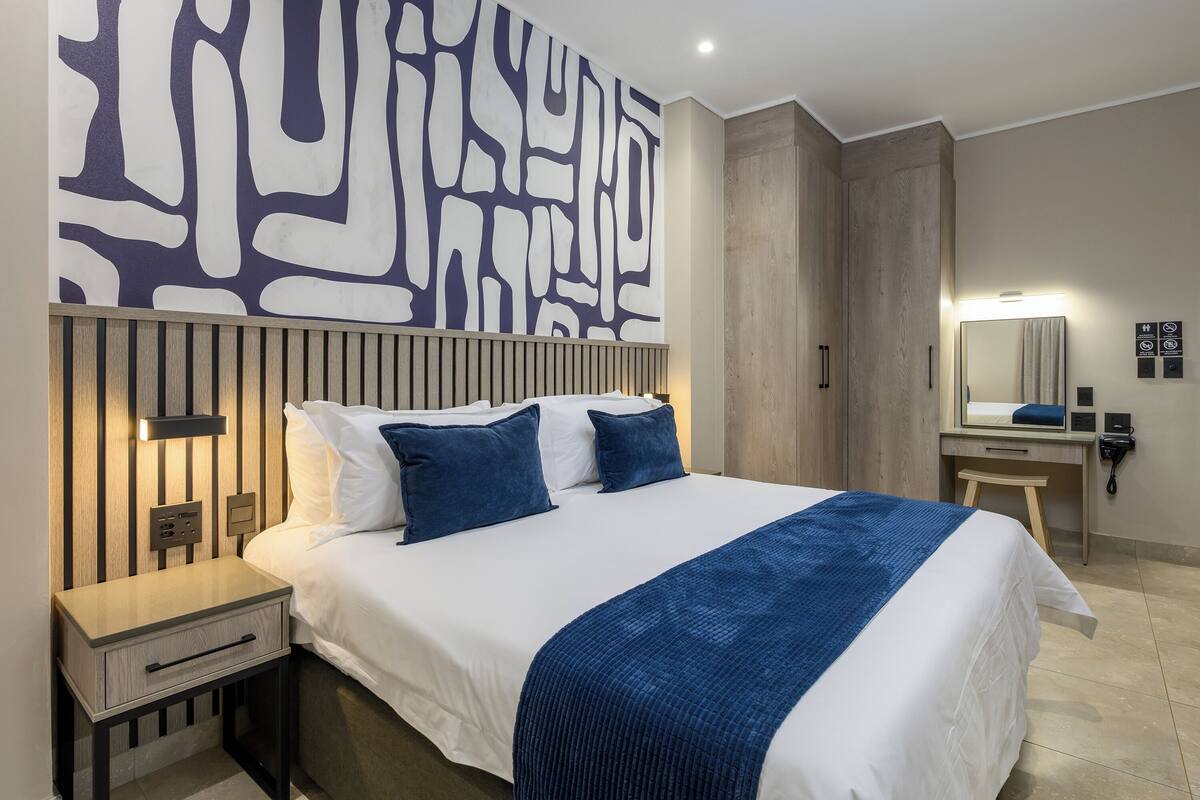 A hotel room with a bed, bedside table, wall art, a mirror, and a wooden panel wall.