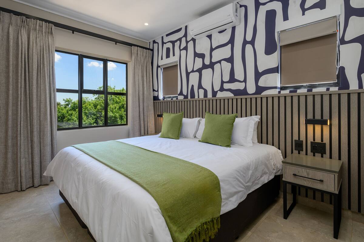 A bedroom with a large bed, a nightstand, a window with curtains, and a wall with a geometric pattern.