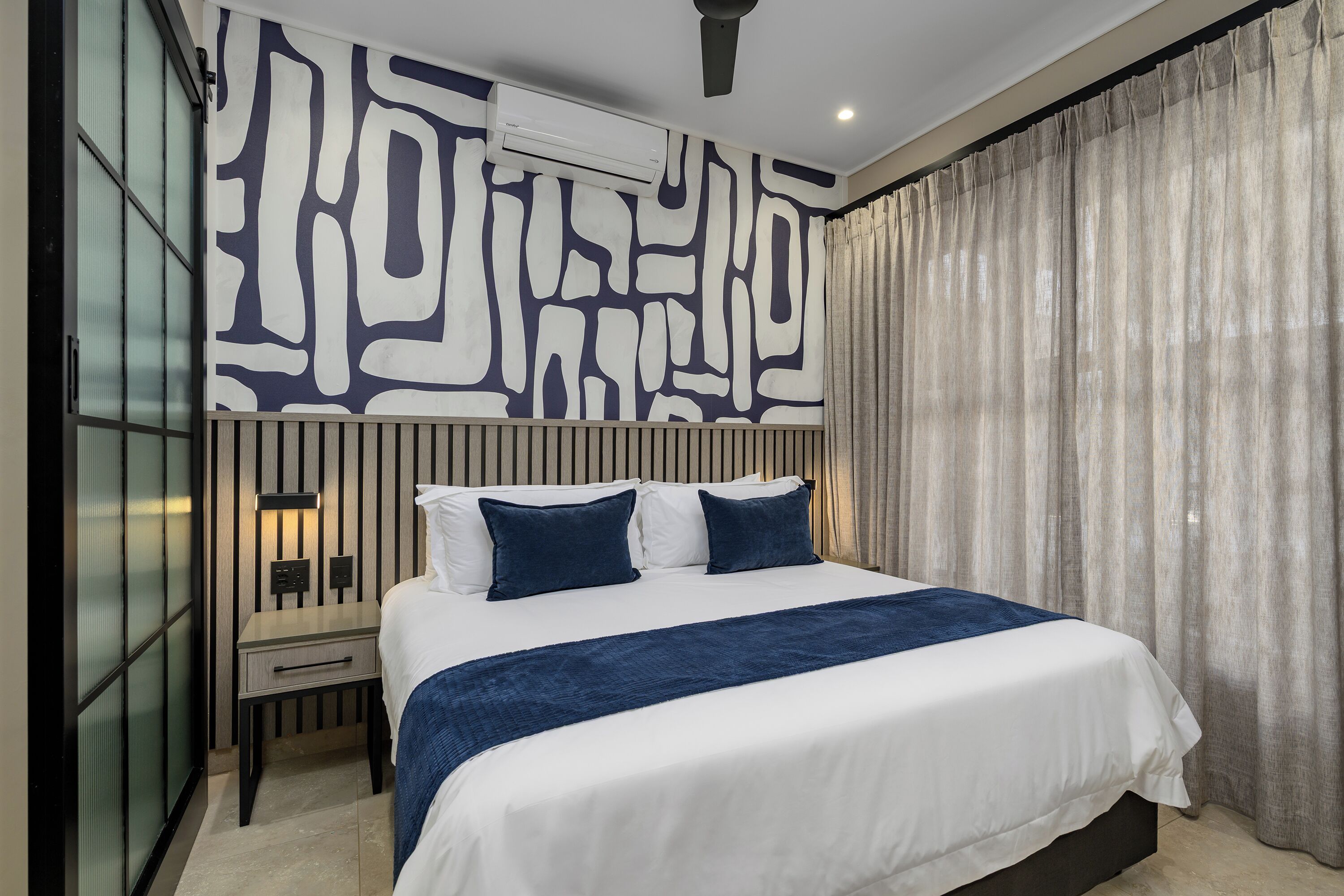 A modern hotel room with a large bed, a nightstand, a wall-mounted air conditioner, and a window with curtains.