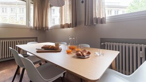 Ein Esstisch mit Croissants, Saft und einer Karaffe, in einem Raum mit weißen Stühlen und Blick auf Gebäude durch große Fenster.