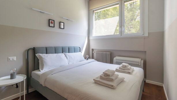Uma cama bem arrumada com roupa de cama branca e toalhas dobradas, um painel de cabeceira estofado cinza, uma mesa de cabeceira com abajur e uma janela com vista para vegetação.