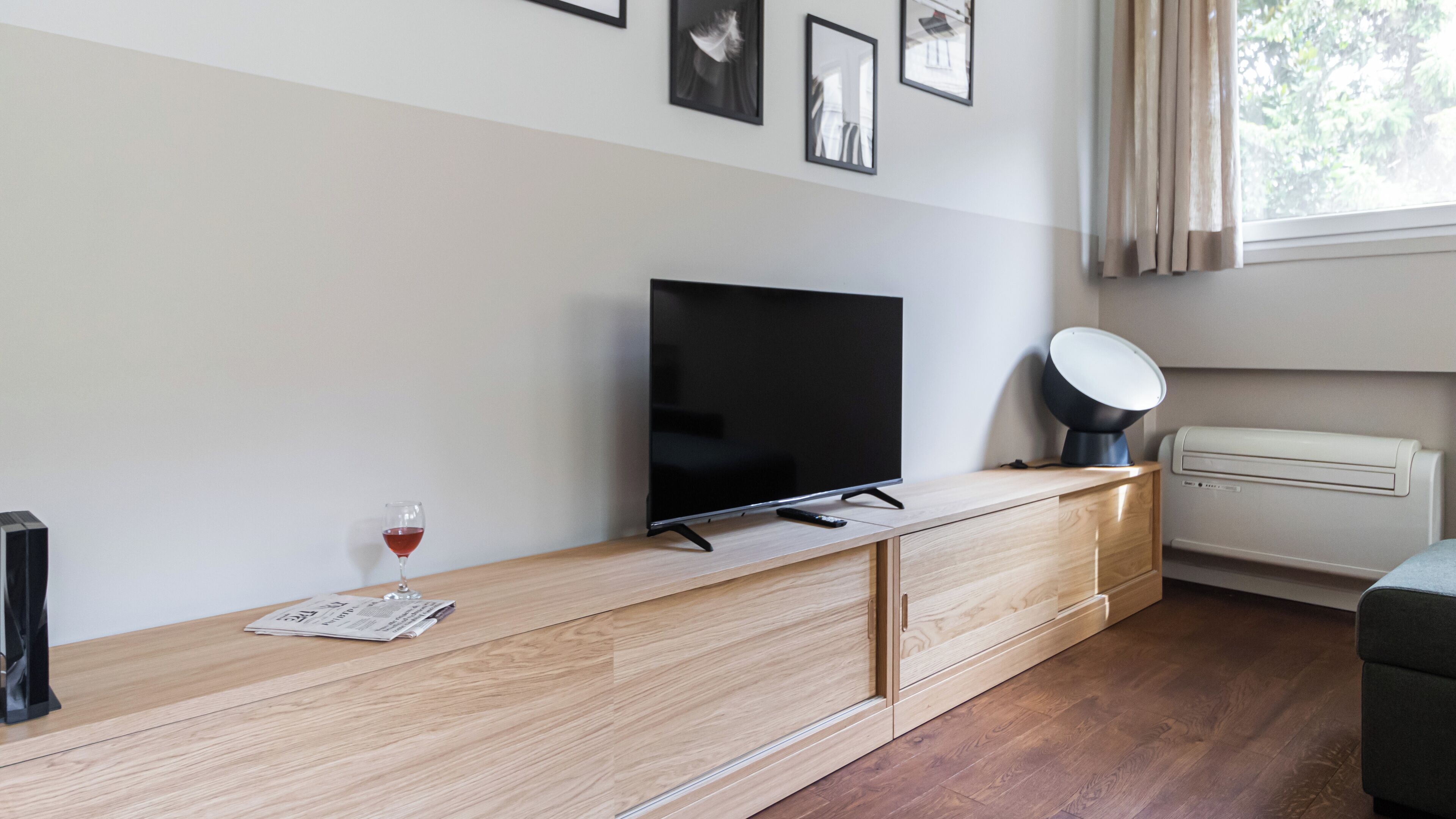 A modern living room with a flat-screen TV on a wooden console, a remote control, a glass of red wine, and a ceiling light fixture.