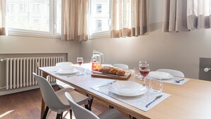 Uma sala de jantar com uma mesa comprida, cadeiras brancas e um radiador. Há pratos, copos e uma cesta de pão sobre a mesa.