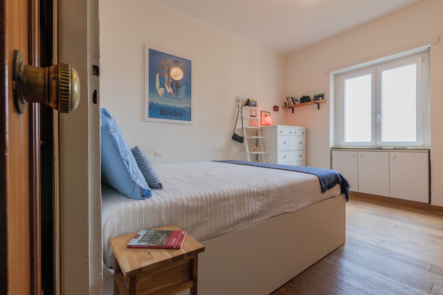 Une chambre à coucher avec un lit, une table de chevet sur laquelle se trouve un livre, une commode, une échelle et une fenêtre.