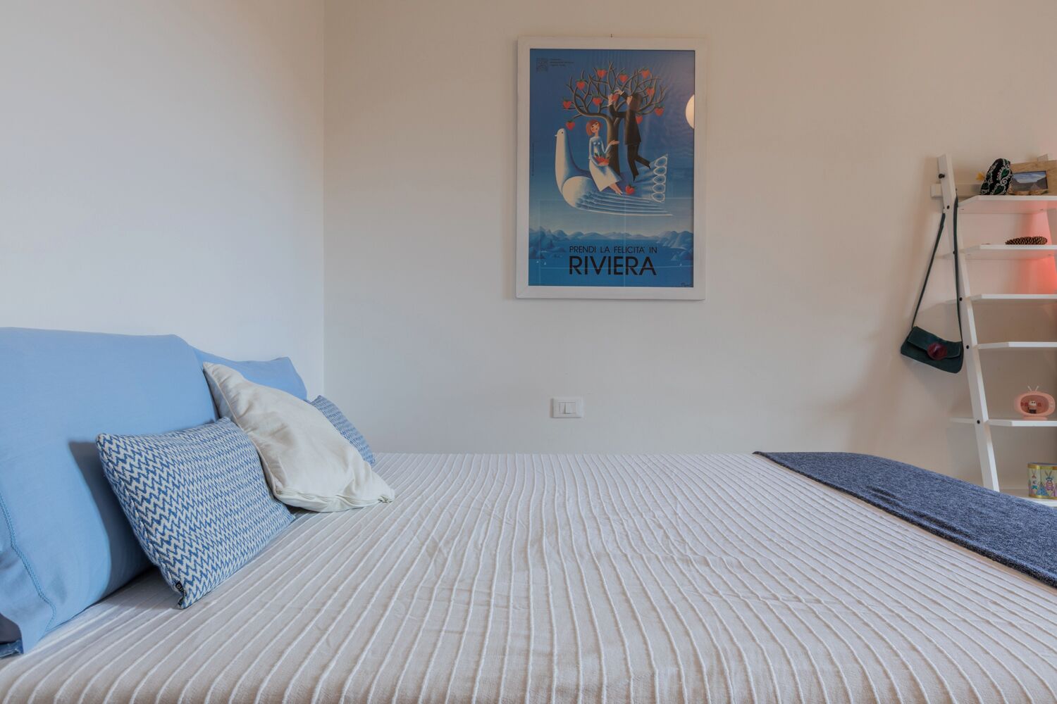 Een slaapkamer met een bed, kussens, een poster aan de muur en een witte plank met spullen.