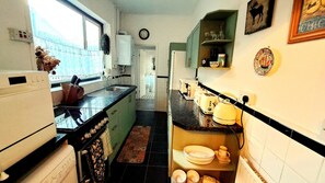 Eine schmale Küche mit Kühlschrank, Spüle und Mikrowelle. Ein Fenster mit Vorhängen, eine Uhr an der Wand und eine Tür zu einem anderen Raum.