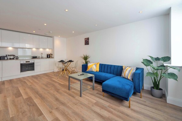 A modern living room with a blue sofa, a glass coffee table, and a dining area with a table and chairs. - Contemporary & Bright 1 Bed Apartment in Gravesend (Gravesend)