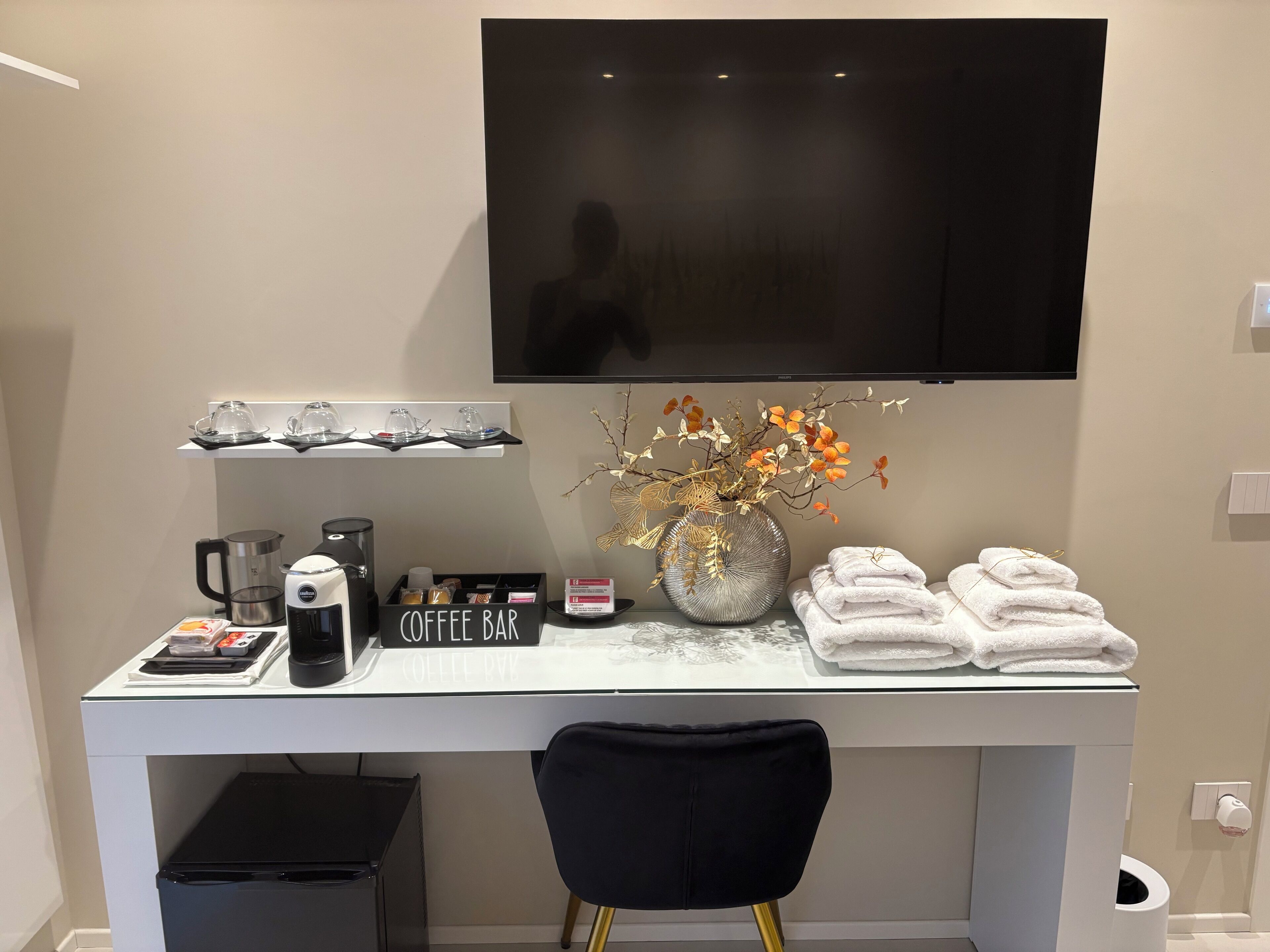 A modern hotel room with a coffee bar, a flat-screen TV, a vase with dried flowers, and neatly folded towels.