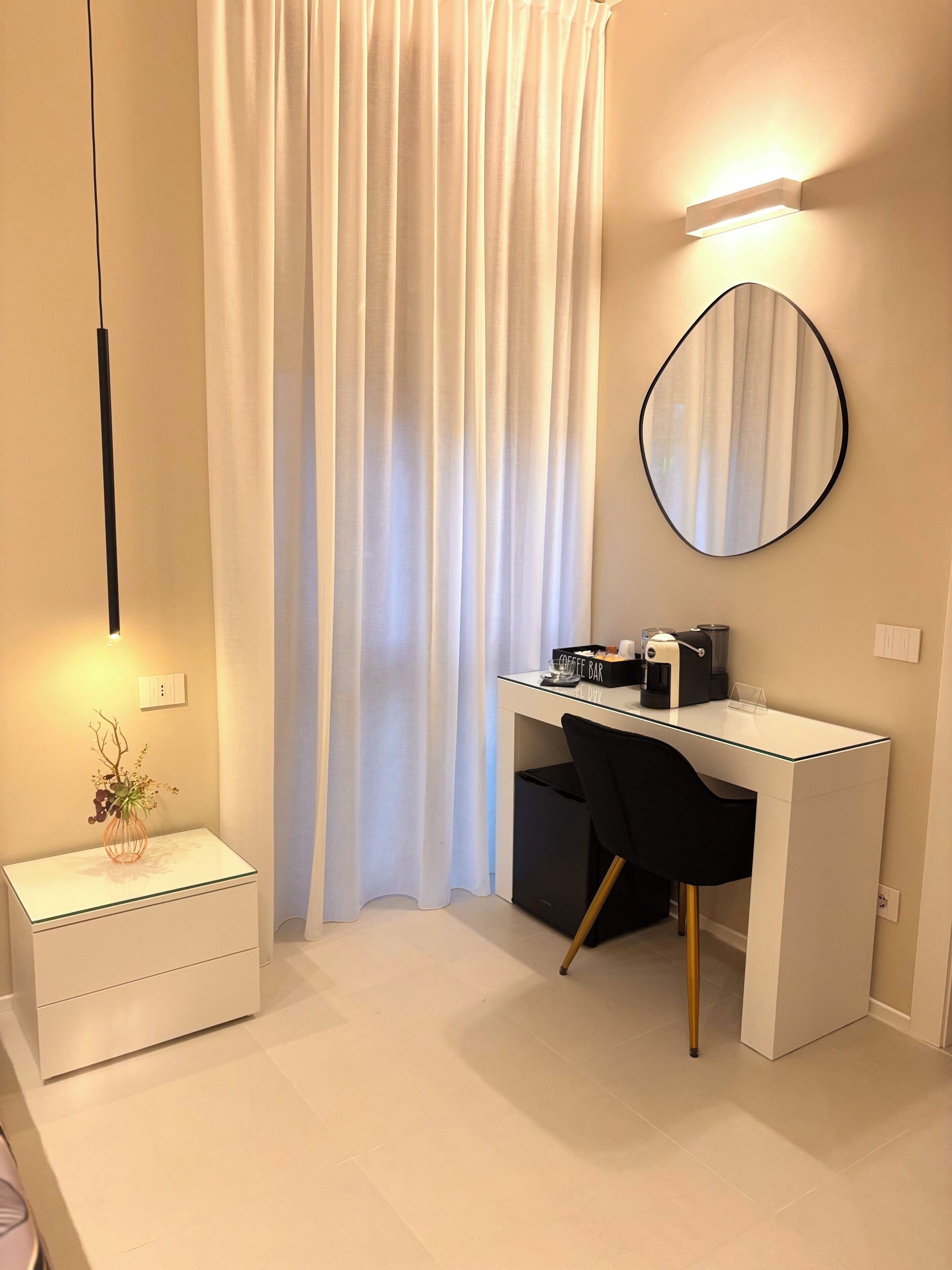 Un dormitorio moderno con un escritorio blanco, una silla negra, una mesita de noche con un florero y un espejo grande en la pared.