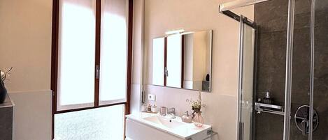 A modern bathroom with a large sink, a separate toilet, a glass shower enclosure, and a large window with blinds.