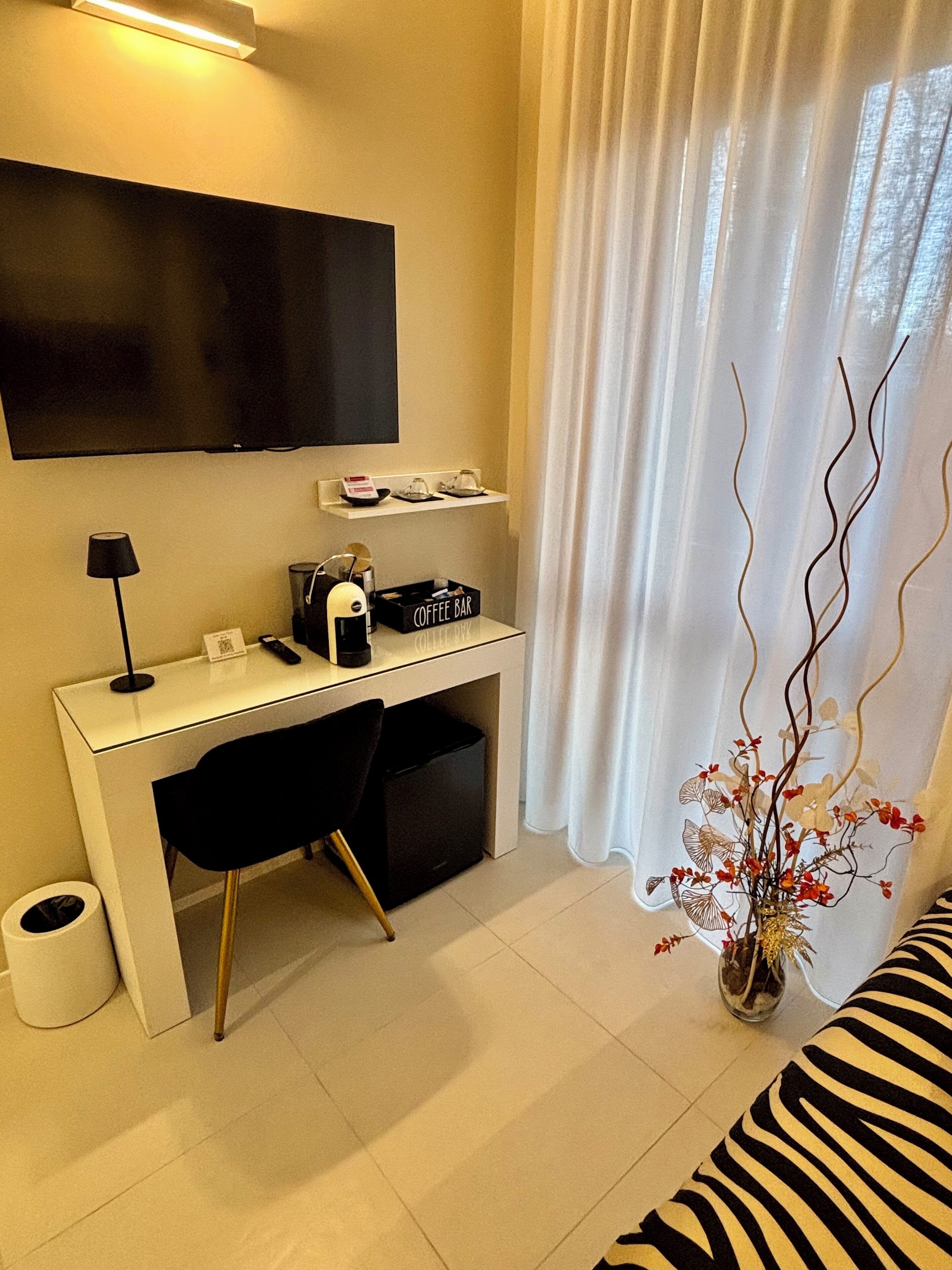 Een moderne hotelkamer met een wit bureau, een flatscreen televisie, een koffiehoekje, een zwarte stoel, een prullenbak en een sierplant.