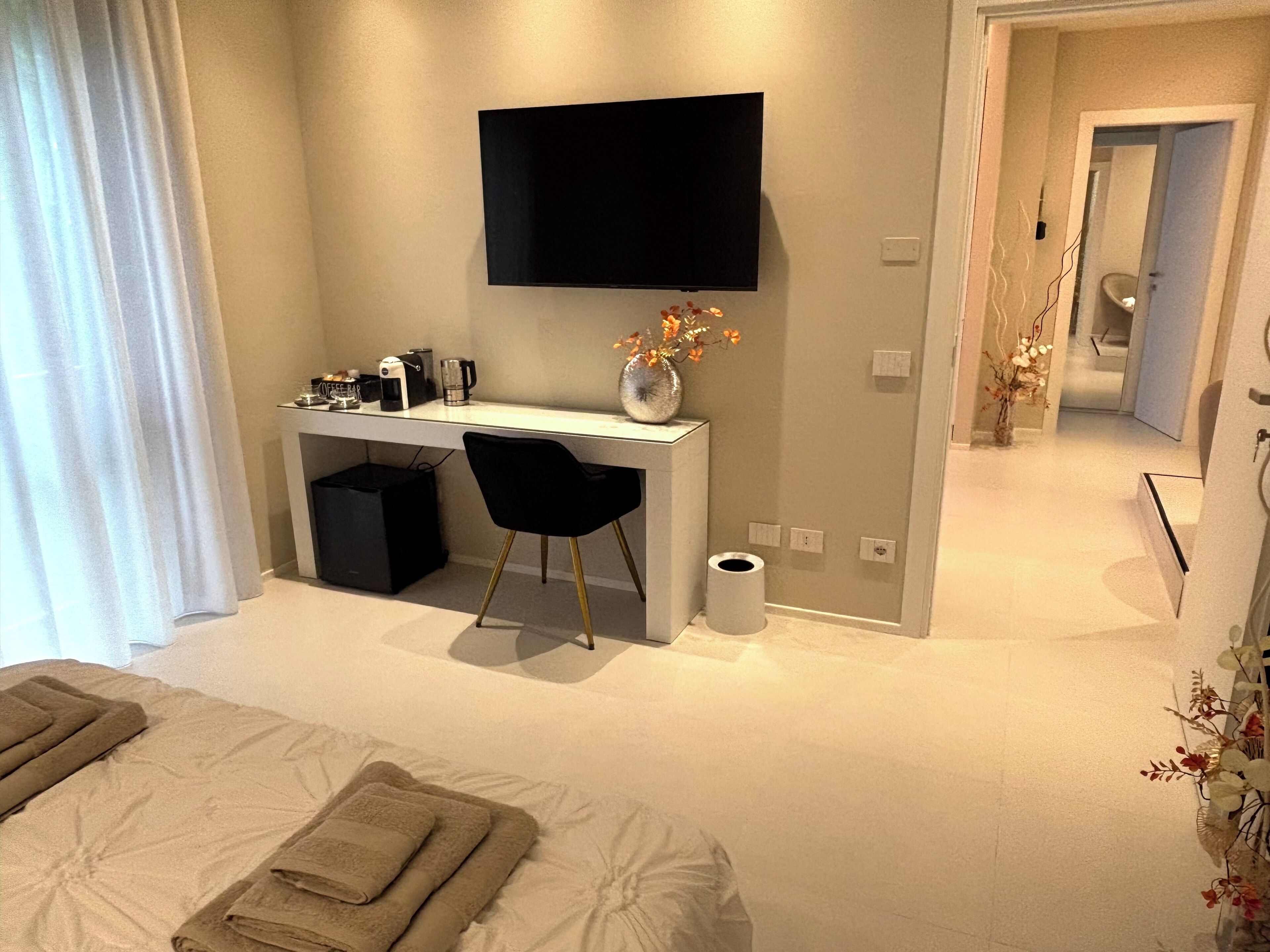 A modern hotel room with a bed, a desk with a chair, a flat-screen TV, a vase with flowers, and a toilet.