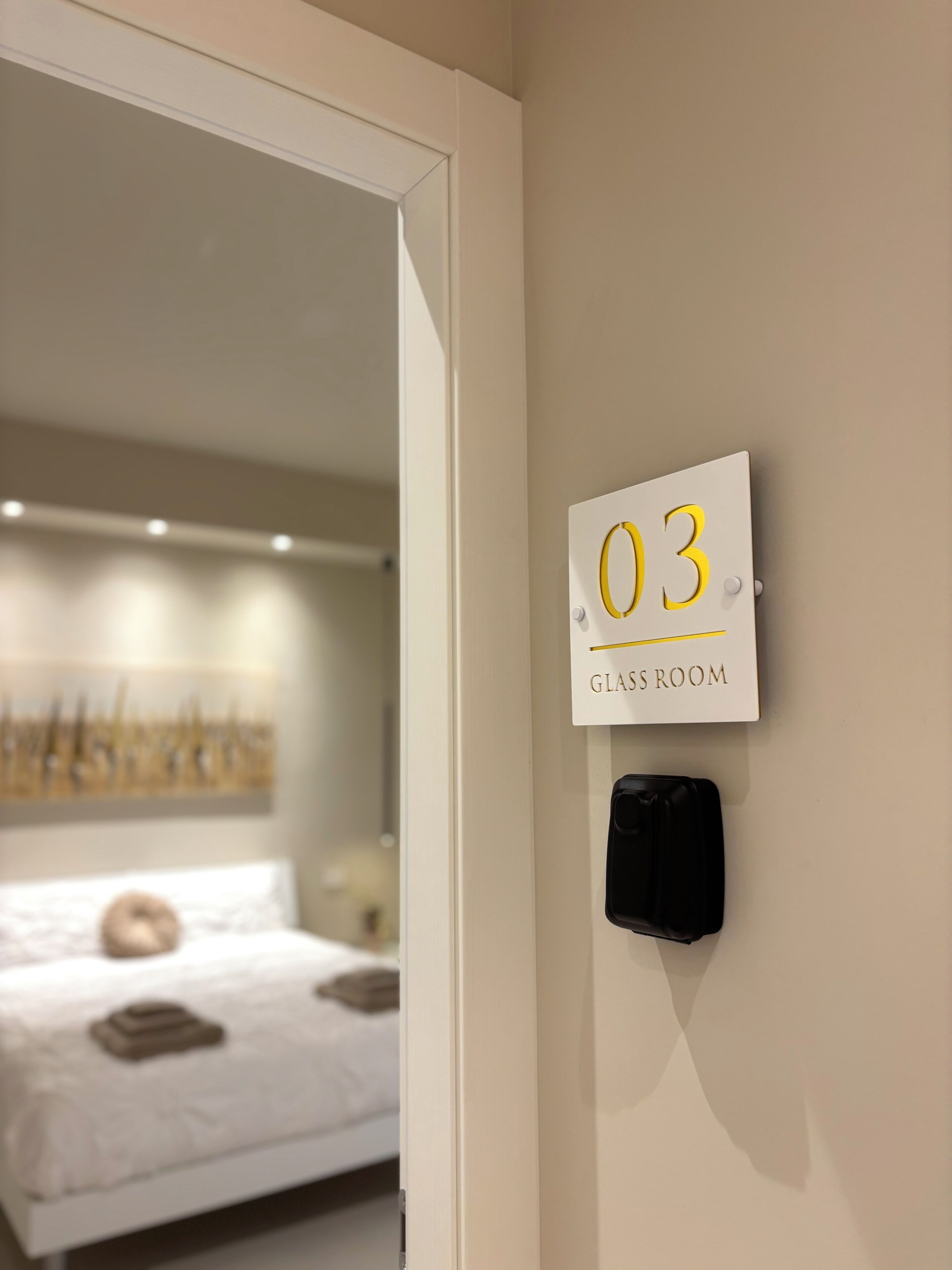 Une chambre d’hôtel avec un lit, une plaque fixée au mur indiquant le numéro de chambre 03, et un objet noir sur le mur.