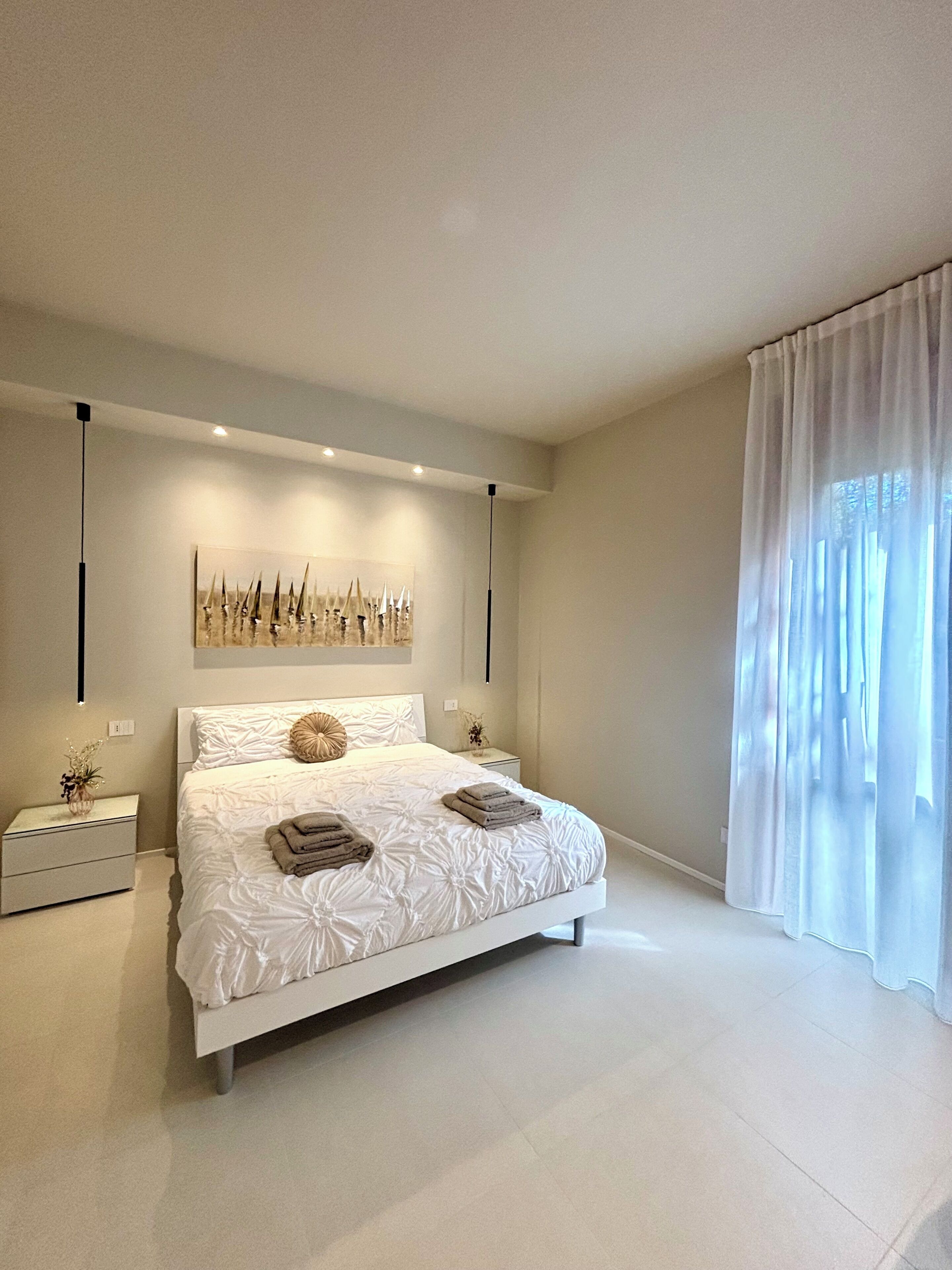 Una camera da letto moderna con un letto grande, comodini, un'opera d'arte appesa al muro e un'ampia finestra con tende leggere.