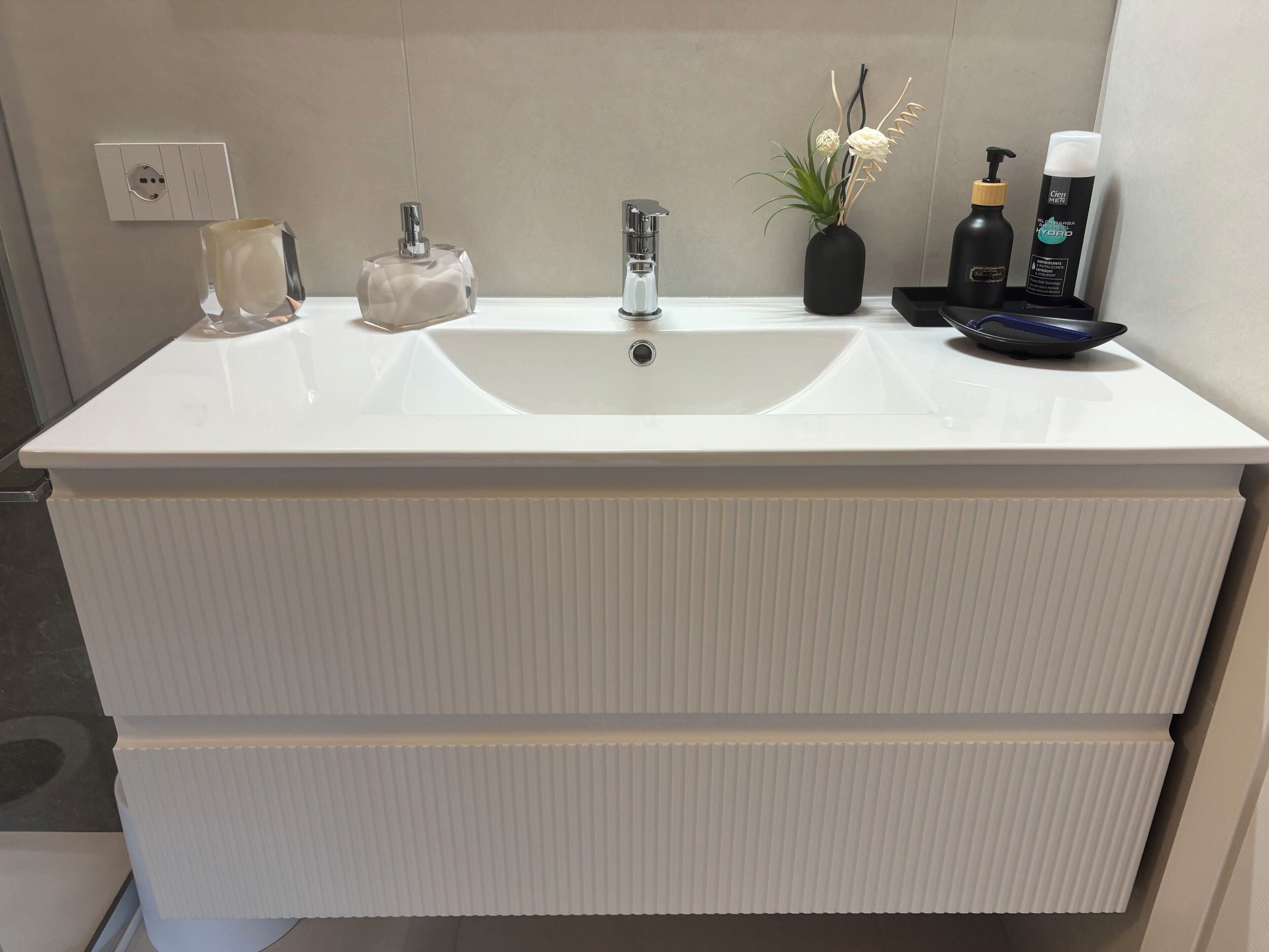 A bathroom with a large white sink, a mirror, a white towel, a glass soap dispenser, a black bottle, and a small potted plant.