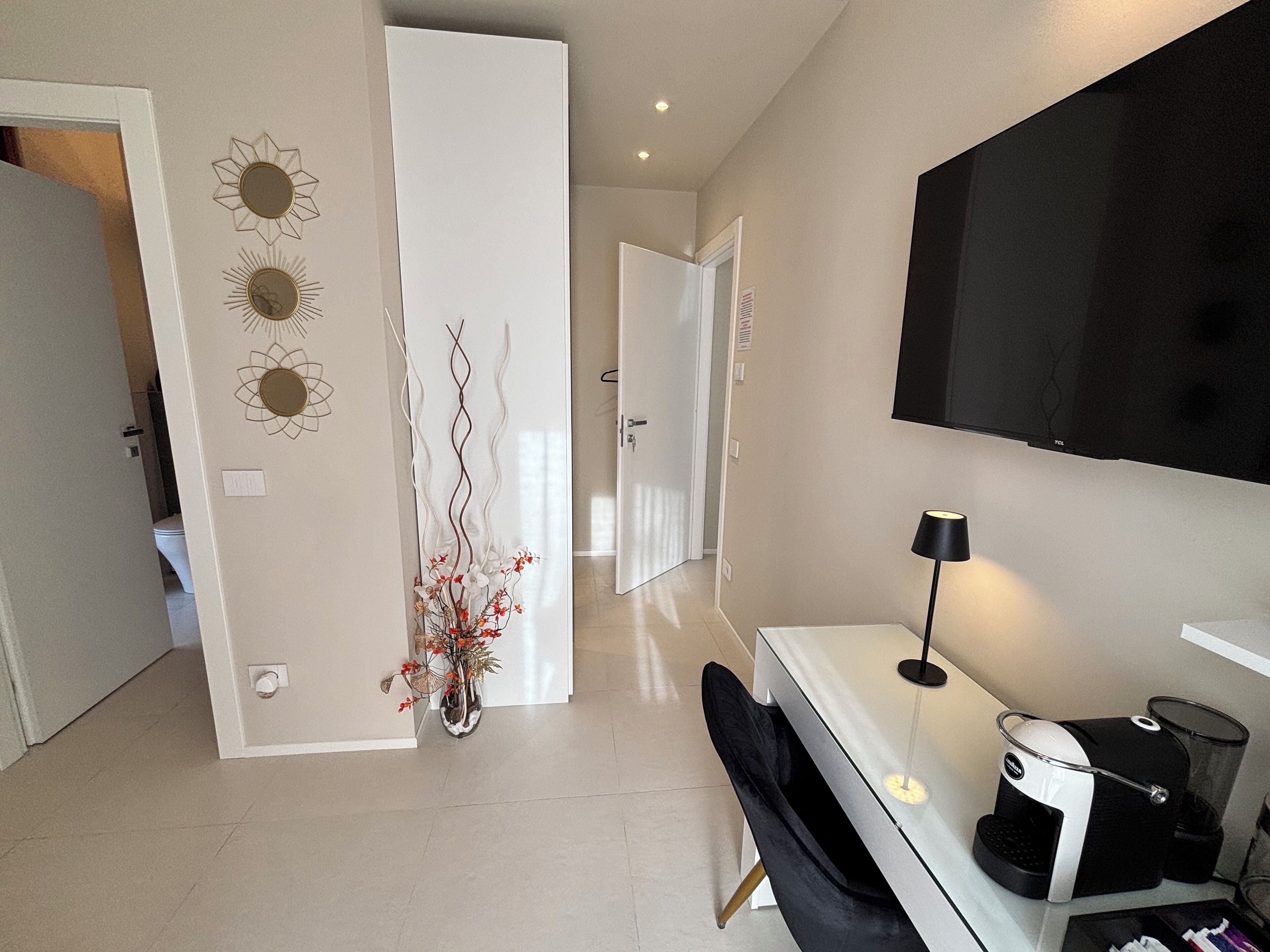 Quarto de hotel moderno com escrivaninha branca, cadeira preta, cafeteira e televisão fixada na parede.