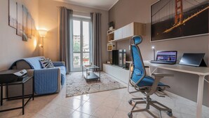 Een moderne woonkamer met een bank, een bureau met een laptop, een stoel, een televisie en een groot raam met gordijnen.