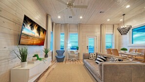 A modern living room with a wooden floor, a large TV mounted on the wall, a sofa, a desk with a chair, and a dining area with a table and chairs. - 2 Homes, Sleeps 22 | 2 Private Pools, Firepit, Steps to Beach Boardwalk Access (Port Aransas)