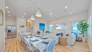 A modern kitchen with a stainless steel refrigerator, a dining area with a long table and blue-striped chairs, and a living area with a sofa and a large window. - 2 Homes, Sleeps 22 | 2 Private Pools, Firepit, Steps to Beach Boardwalk Access (Port Aransas)