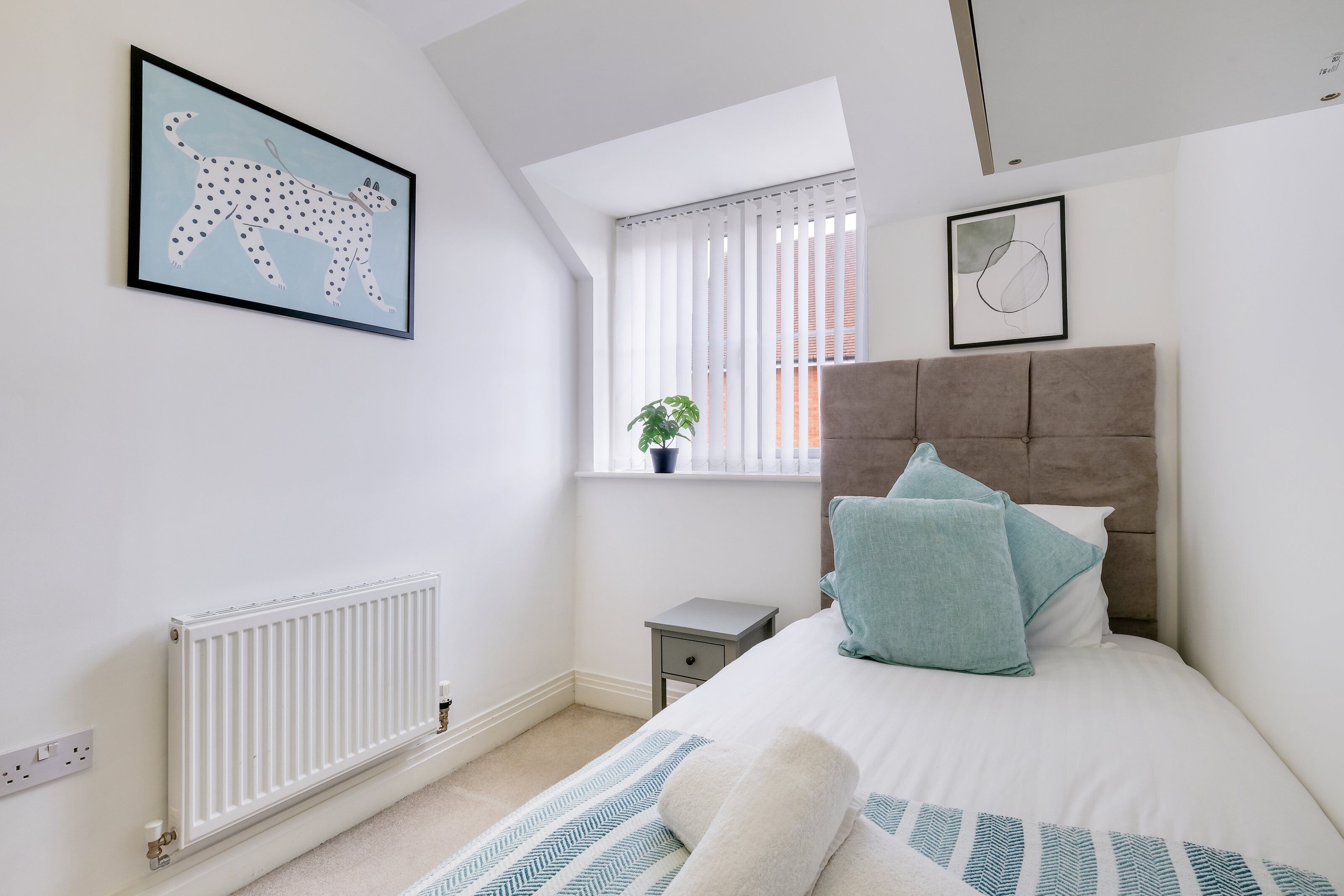 A bedroom with a bed, a radiator, a window with blinds, a small bedside table, and a framed picture on the wall.