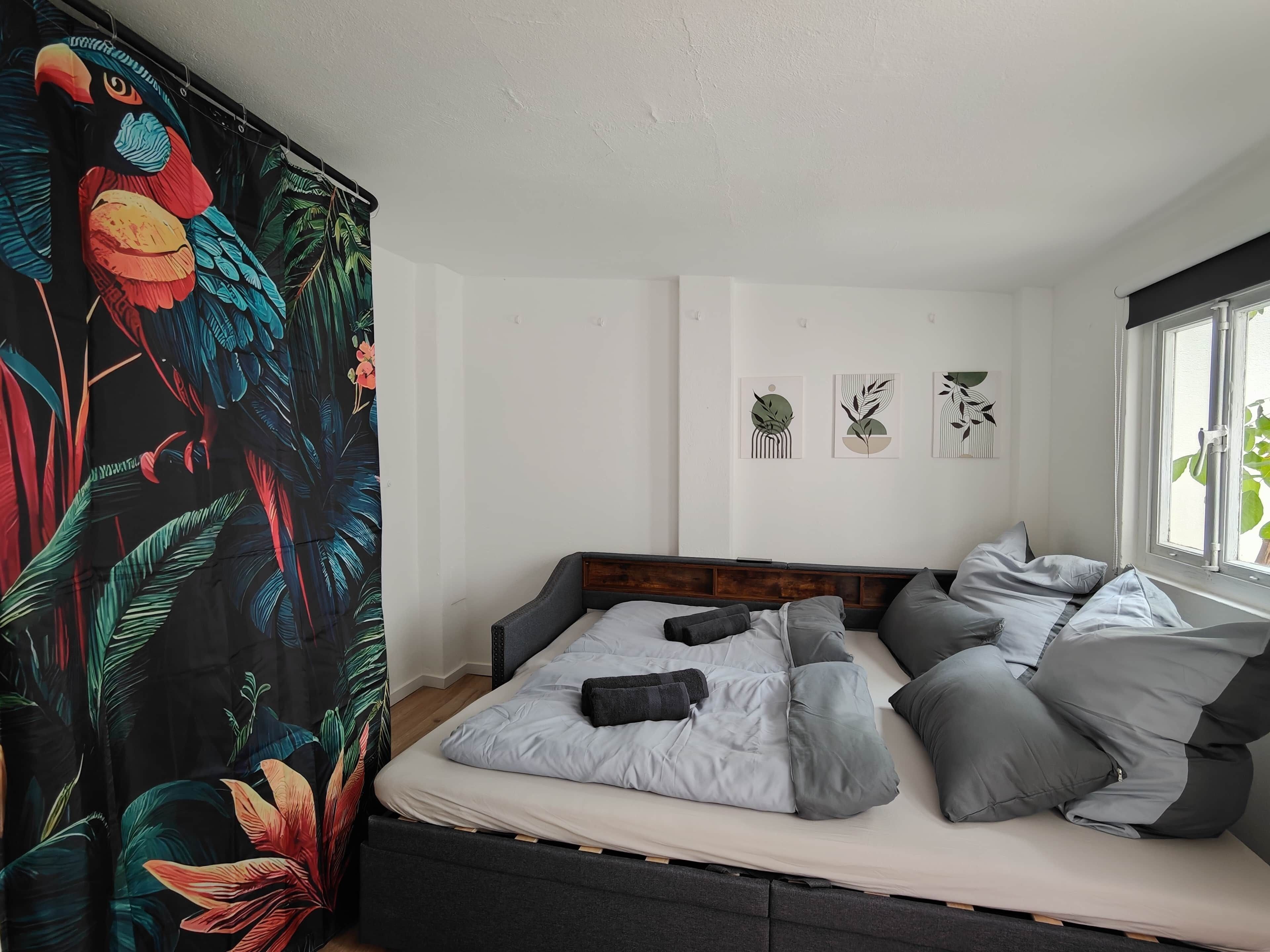 Een slaapkamer met een bed, grijs beddengoed en een gordijn met tropische motieven.