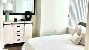 A bedroom with a large bed, a dresser with drawers, a mirror, and a window with blinds. - Beautiful Two-Bedroom Escape Ocean & City Views from Every Room (San Diego)