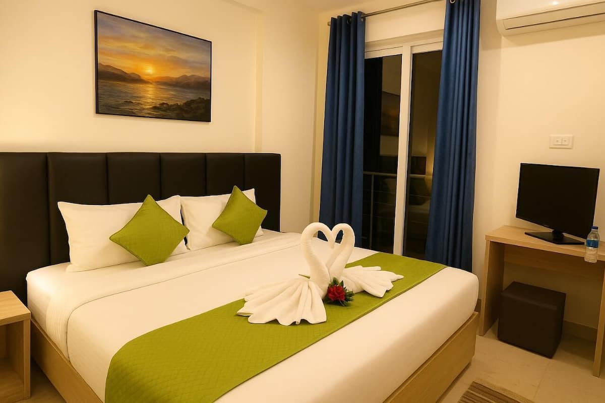 A neatly made bed with white linens, green pillows, and a green bedspread. There is a swan-shaped towel arrangement on the bed, and a painting of a sunset on the wall.
