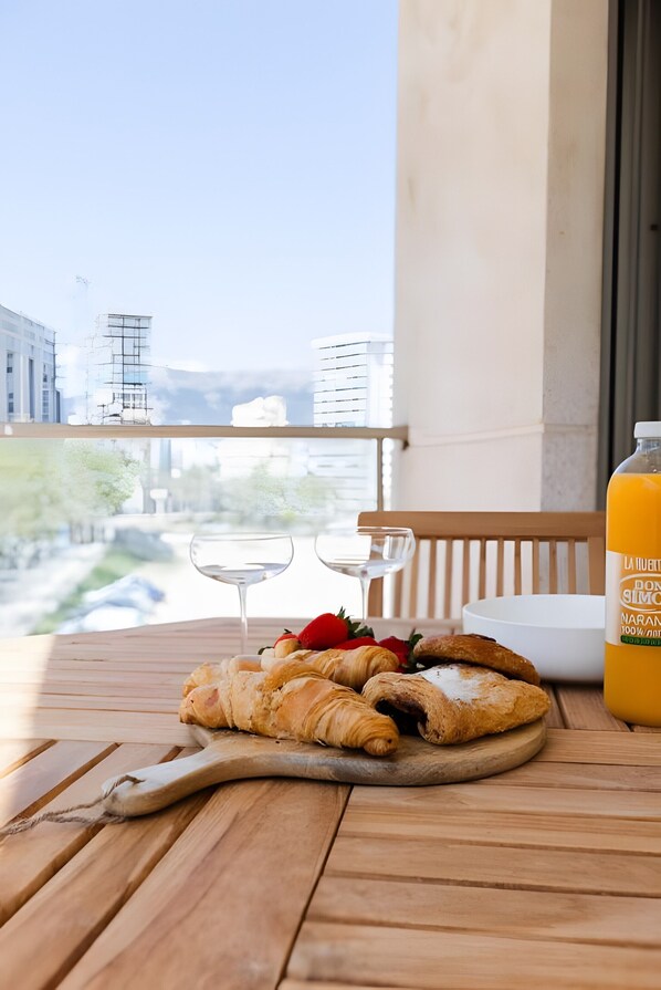 A balcony with a wooden table, a bottle of orange juice, croissants, strawberries, and a glass of water.