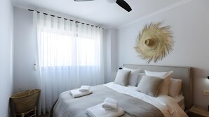 Ett hotellrum med en säng, en takfläkt, ett stort fönster med gardiner, en dekorativ detalj som hänger på väggen och ett nattduksbord.