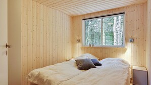 A bedroom with a wooden paneled wall, a window with a view of trees, a bed with pillows, and a bedside table.