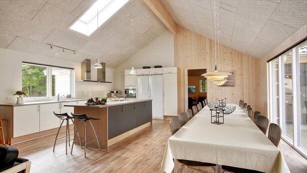 A modern kitchen with a central island, a dining area with a long table, and a view through large windows.