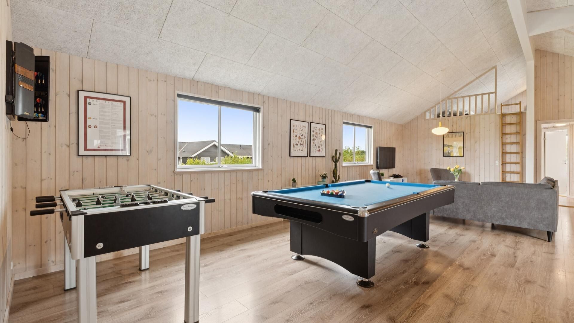A room with a foosball table, pool table, wooden floors, and a view of houses outside the window.