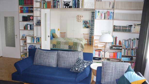 A room with a blue sofa, a bed with a green quilt, and a bookshelf filled with books.