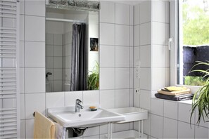A bathroom with white tiles, a sink, a mirror, a shower, and a window with a view of greenery.