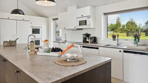 A modern kitchen with white cabinets, a large island, a microwave, and a window with a view of trees. - VillaVilla 598 - Rømø, Sydjylland (Rømø)