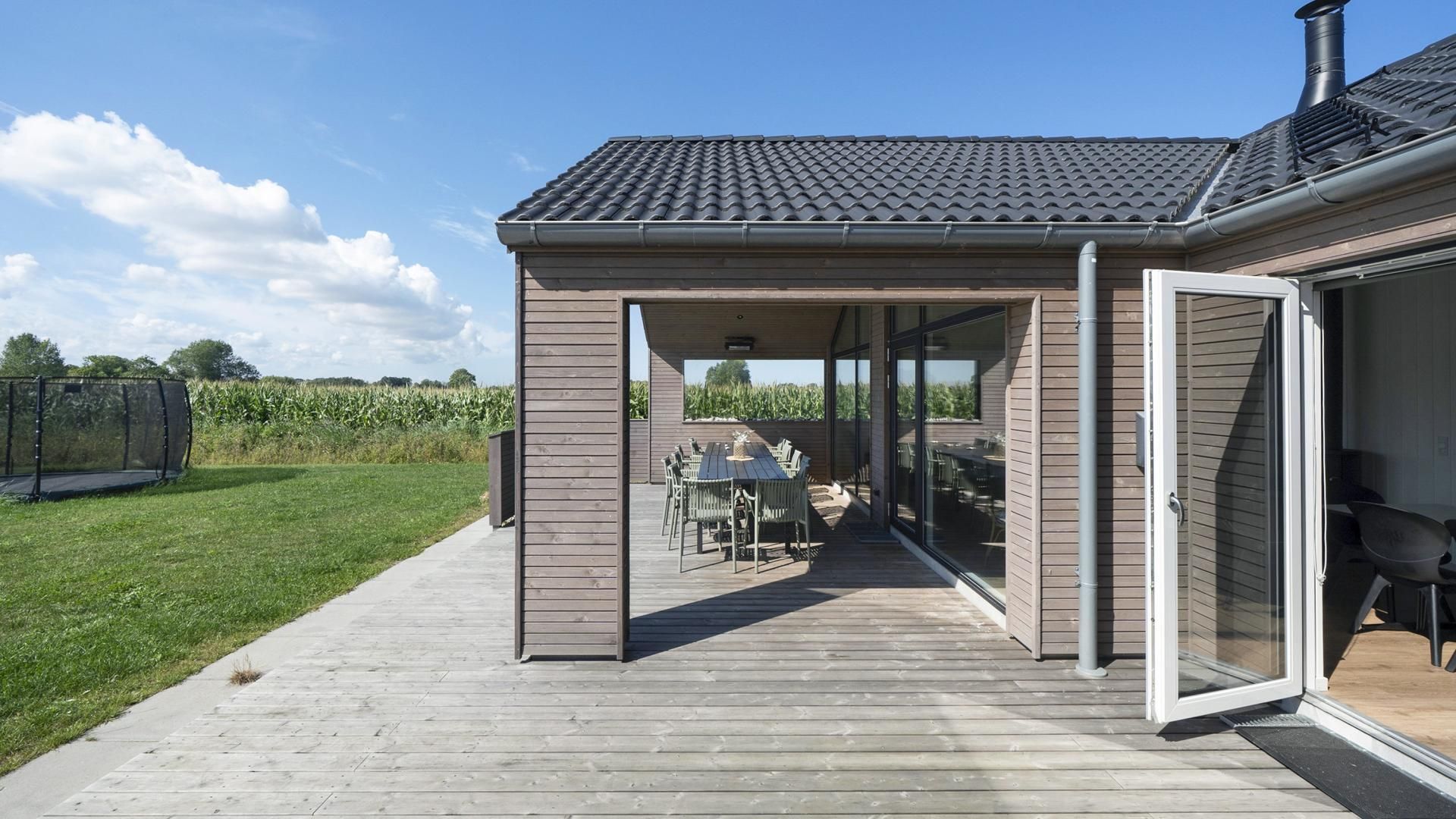 A covered outdoor patio with a table and chairs, a wooden deck, and a view of a cornfield.