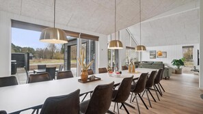 A modern dining area with a long table, multiple chairs, pendant lights, and a view of the outdoors. - VillaVilla 621 - Dueodde, Bornholm (Aakrikeby)
