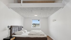 Een lichte, moderne slaapkamer met een bed, nachtkastje, lamp en een raam met uitzicht op gebouwen.