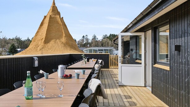 Een houten terras met een lange eettafel, een zandduin op de achtergrond en een modern gebouw met grote ramen.