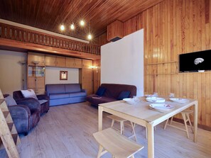 Ein gemütliches Blockhausschlafzimmer mit Holzbalkendecke, einem Esstisch gedeckt mit Tellern und Gläsern, einer Couch und einem Fernseher, der den Mond zeigt.