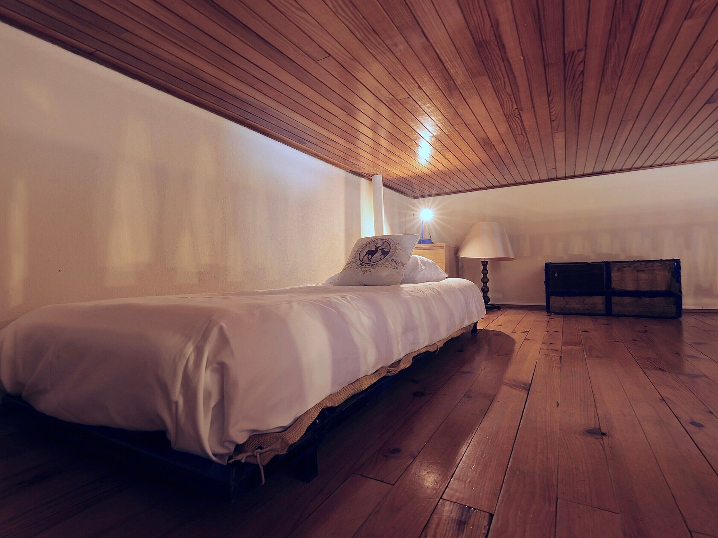 Ein Schlafzimmer mit Holzbalkendecke, einem Einzelbett, einem Nachttisch, einer Lampe und einer Holzkiste.