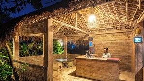 A wooden outdoor bar area with a thatched roof, a wooden counter, and a person seated behind it.