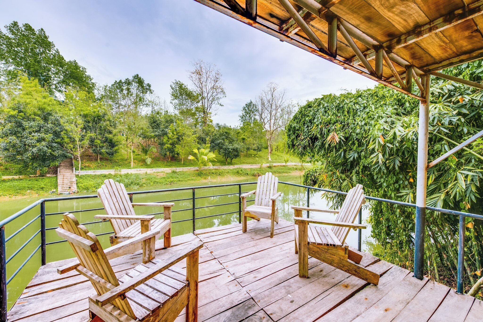 A wooden deck with four Adirondack chairs overlooking a river, surrounded by lush greenery.