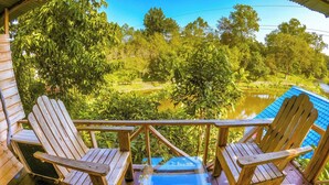 A wooden deck with two Adirondack chairs and a glass table, overlooking a pond and lush greenery.