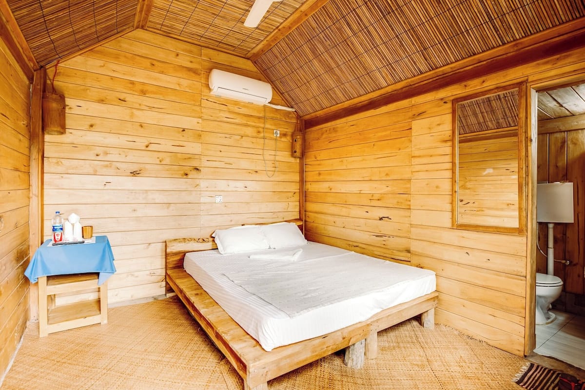 A wooden cabin room with a bed, a small table with a blue tablecloth, and a window.