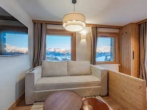 A cozy room with a sofa, a round wooden table, and a large window offering a mountain view.