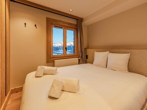 A neatly made bed with two pillows and two rolled towels, a wooden headboard, a window with a view of snow-capped mountains, and a radiator under the window.