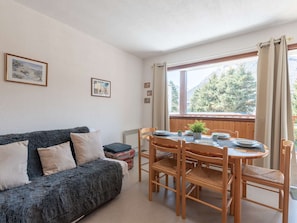 A cozy room with a dark blue sofa, a wooden dining table with chairs, and a window with curtains. - Cozy duplex for 4 people, near shops and ski slopes (Saint-Lary-Soulan)