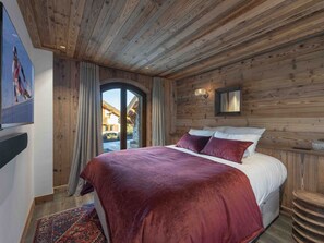 Une chambre à coucher aux murs lambrissés, avec un lit, des rideaux, une lampe fixée au mur et une vue sur l’extérieur à travers une porte vitrée.
